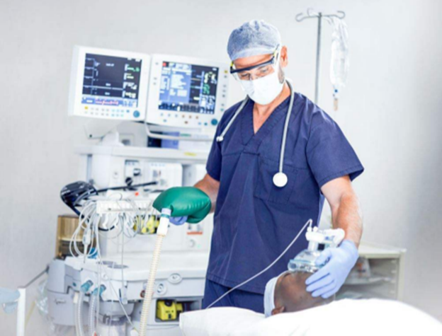 A doctor in scrubs and a mask applies anesthesia to a patient before vascular surgery.
